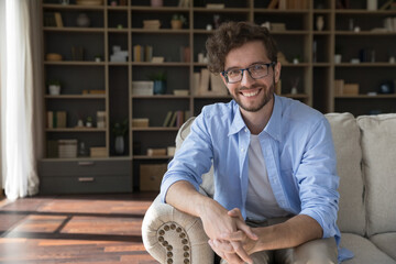 Happy handsome young 20s business man in glasses sitting on couch at home with bookshelves in...