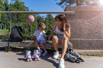 Mom putting on inline skates rollers to daughter child in public park in summer. Family leisure...