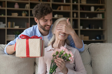 Cheerful adult son guy giving surprise gift to old senior mom, covering woman eyes with hand,...