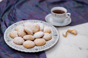 Kahk with sugar sprinkled on top, in a rounded china plate and a cup of tea, on white marble background, (Kahk is a traditional cookies after Ramadan for eid)

