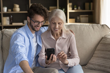 Happy adult child man and senior older mom using mobile phone at home together, talking on video...