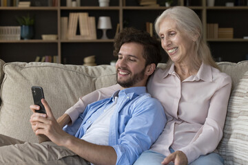 Positive senior mom and adult child guy using mobile phone for family wireless connection on Internet, talking online video conference call, resting on sofa at home, holding gadget, smiling, laughing