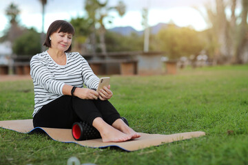 Fototapeta premium World health day. Smiling adult woman in casual sportswear sitting on yoga mat, using cellphone and relaxing to listen music with headphones after training with massage roller on green grass outdoors