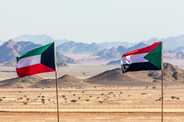 Kuwaiti and Sudanese flags waving togetner on the wind in Saudi Arabian desertwith mountains in...