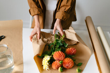 Obraz na płótnie Canvas A young woman wraps a bouquet of orange dahlias in kraft paper for a gift. Flower shop, small business and flower delivery concept.