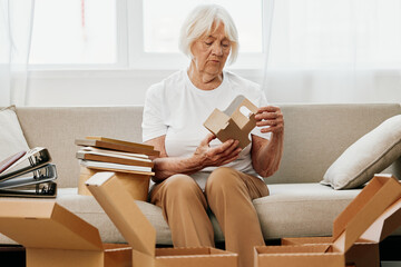 elderly woman sits on a sofa at home with boxes. collecting things with memories albums with photos and photo frames moving to a new place cleaning things and a happy smile. Lifestyle retirement.