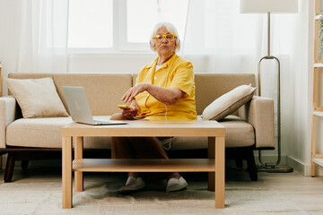 Happy elderly woman with laptop typing in headphones smile sitting at home video call, bright modern interior, lifestyle online communication.