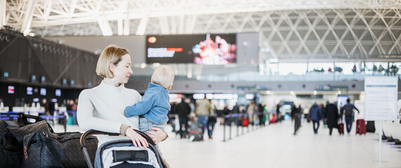 Obraz premium Motherat travelling with his infant baby boy child, walking, pushing baby stroller and luggage cart at airport terminal station. Travel with child concept