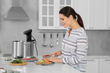 Woman cutting carrot near pot with sous vide cooker at table in kitchen. Thermal immersion...