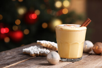 Tasty eggnog with cinnamon, cookies and baubles on wooden table against blurred festive lights. Space for text