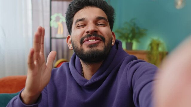 POV of indian man blogger taking selfie on smartphone, communicating video call online with social media subscribers followers, recording vlog stories. Young hindu guy at home apartment room on couch