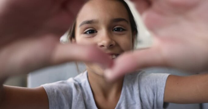 Close up pre-teen girl smile staring at camera through joined fingers showing symbol of love, heart shape, feel happy advertise professional dental services, braces need, looking overjoyed. Affection