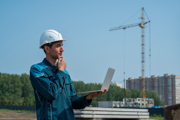A male builder in a helmet stands against the background of a construction site with a laptop in his hands. 