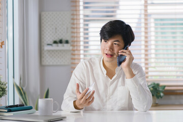 A millennial Asian male freelancer is talking on his smartphone while working remotely from home office with his laptop.