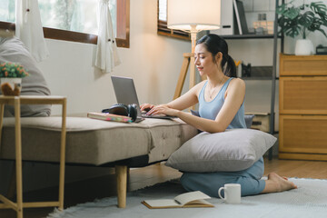 Millennial Asian girl sits on sofa in living room, studying on laptop making notes.The concentrated young woman works on computer and writes in her notebook, taking online course or training at home.