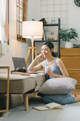 Obraz premium Millennial Asian girl sits on sofa in living room, studying on laptop making notes.The concentrated young woman works on computer and writes in her notebook, taking online course or training at home.