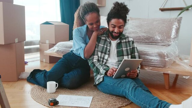 Young multicultural couple African American woman and Arabian man together choose furniture for own new apartment or draw up design project for renovation sit on floor near sofa and cardboard boxes