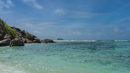 Fototapeta na wymiar Tropical idyll. Calm turquoise ocean and blue sky with clouds. The foam of waves over the reef in the distance. Green vegetation on the rocky slope of the island. Seychelles. Moyenne Island. 