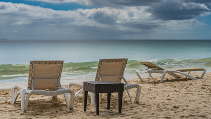 Sun loungers and a wicker table stand on the sandy beach. The waves of the turquoise ocean roll...