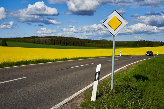 Traffic sign on the side of the road. Yellow rapeseed (Brassica napus) field behind the roadway. Green forest in front of blue cloudy sky. Driving vehicle.