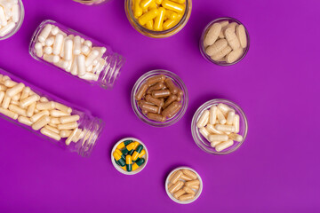 Many small glass bottles and bowls filled with different vitamins and minerals in form of capsules, tablets and pills from above on violet background. Vitamin b, d3, c, a, e, magnesium, zinc.