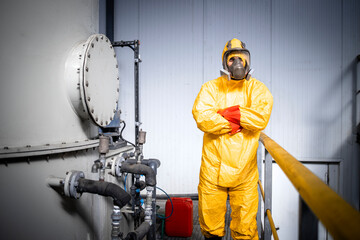 Naklejka premium Portrait of factory worker in protective yellow suit, gas mask and gloves holding arms crossed inside chemicals production plant.
