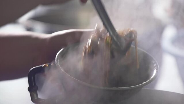 The chef is scooping stir-fried Hokkien noodles in a hot pan with steam into a bowl and wrapping paper for take-away, a traditional dish of Phuket.