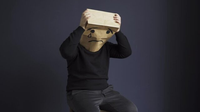 A sad man in a black turtleneck with a bag on his head, with a drawn crying emoticon, holds his head and cries. Emotions and gestures.
