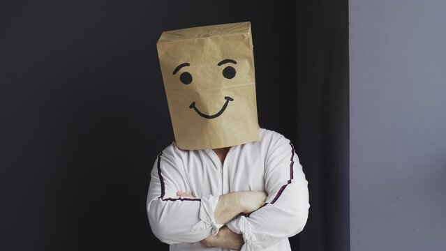 A man in a white shirt with a paper bag on his head, with a painted smiley face, stands against the wall, overwhelmed with feelings, shows a class gesture, with his hand.