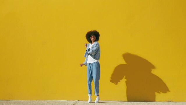 Playful happy young African American woman wear stylish blue denim clothes dancing and jumping funky black teen fashion girl on summer yellow background with copy space. Steady shot, full body