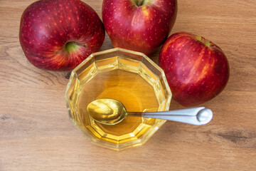 Dessert spoon in a bowl of vinegar and red apples around it, on a wooden background.