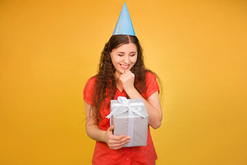 Portrait of a young woman in a paper cap with a gift in her hands isolated on a yellow background, birthday concept.