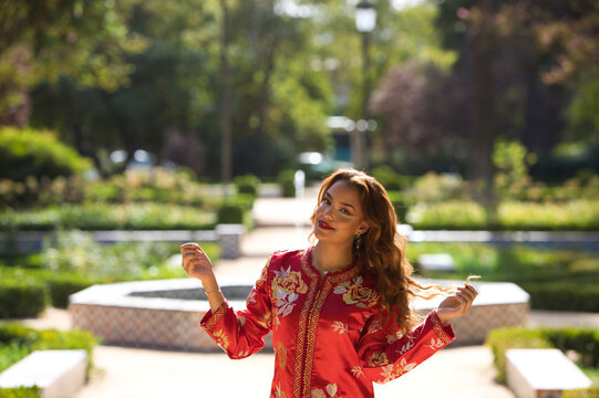 A beautiful woman wears a traditional Moroccan dress in red and embroidered in gold and silver. The girl raises her arms for wedding photos in a Moroccan style garden with fountains and lush greenery