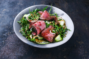 Traditioneller Commonwealth Sonntagsbraten als Aufschnitt Roastbeef mit Zucchini und Zwiebelringen in Rucola Salat serviert als Draufsicht in einer Design Schale 