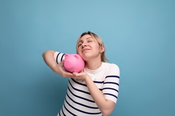 blond attractive young woman in casual attire carefully hugging a piggy bank with savings on a blue background with copy space