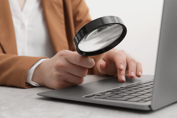 Woman holding magnifier near laptop at light gray table, closeup. Online searching concept