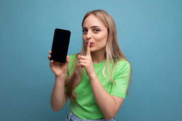 bright blond girl in casual outfit holding a smartphone vertically with a screen mockup covering her mouth with a finger forward on a blue background