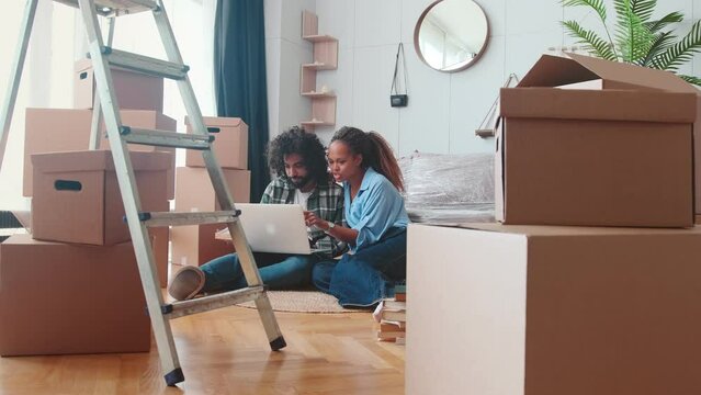 Young cheerful African American woman and Arabian man use laptop to order furniture wanting to freshen up interior after relocation sits on floor in living room with cardboard boxes and stepladder