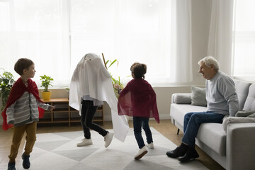 Fototapeta premium Happy senior grandfather watching three boys in homemade super hero costumes chasing each other, running, playing role active game. Grandpa enjoying meeting with grandsons