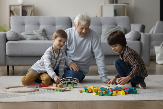 Two cheerful kids and grandpa playing constructor game on heating carpeted floor at home, building city road from mess of toy blocks. Grandkids visiting grandpa, having fun, enjoying leisure