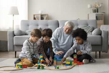 Elderly grandpa watching three multiethnic little grandsons playing game with toy construction blocks on warm floor, building city towers model together, enjoying family playtime, teamwork