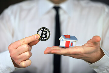 A businessman in a white shirt and black tie holds a bitcoin and miniature house in his hands.