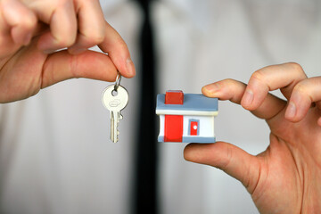 A businessman in a white shirt and black tie holds a keys and miniature house in his hands.
