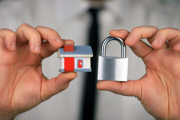 A businessman in a white shirt and black tie holds a silver padlock and miniature house in his hands. Home security.