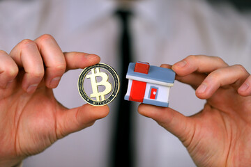 A businessman in a white shirt and black tie holds a bitcoin and miniature house in his hands.