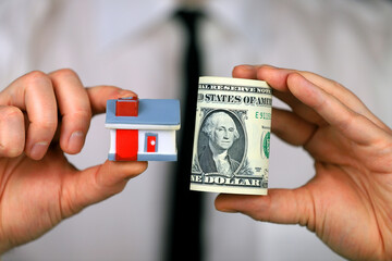 A businessman in a white shirt and black tie holds a bundle of dollars and miniature house in his hands.