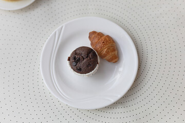 A croissant and a chocolate muffin on a white plate on white dotted background, top view.  Morning snack, copy space, flat lay.
