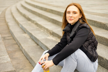 Young woman in black jacket and jeans sit on steps with coffee cup outdoor 

