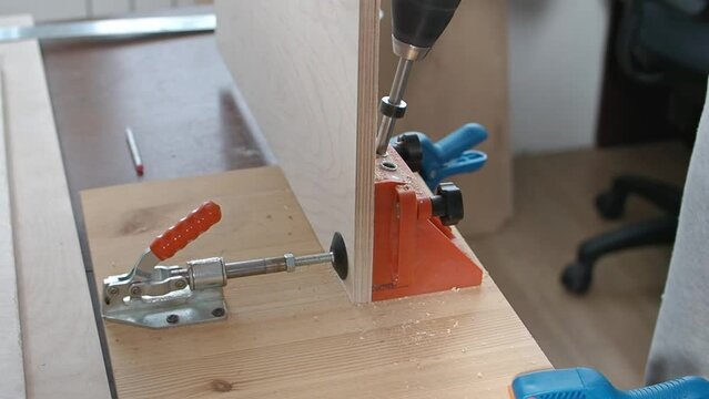 Carpenter caucasian man use pocket hole jig tool and drill bit to make strong joints in sheet of plywood. Furniture part is fixed vertically on the table with special clip