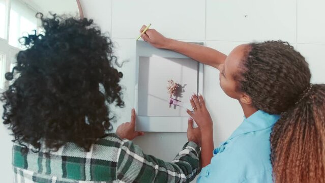 Young friendly African American woman and Arabian man together hang picture on wall marking place with pencil wanting to decorate interior of apartment or office stand in room near window